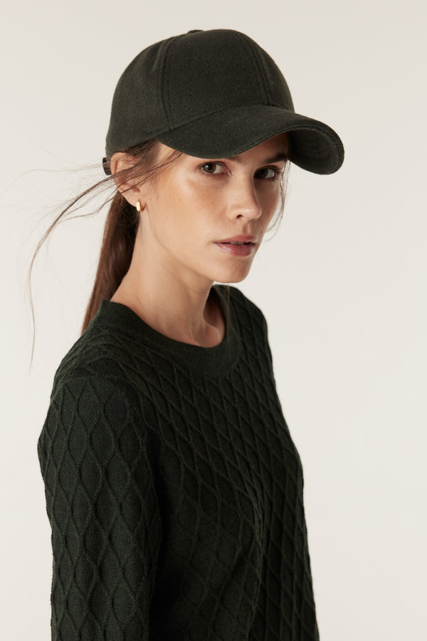Woman wearing a moss green merino wool jumper with diamond cable knit and a matching baseball cap.