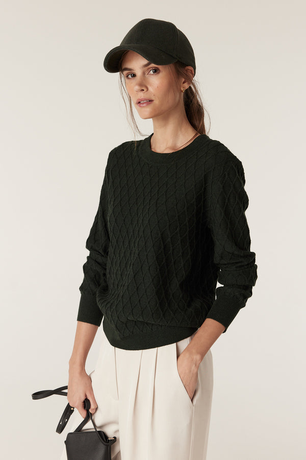 Woman wearing a moss green merino wool jumper with diamond cable knit, crew neck, and rib trims.