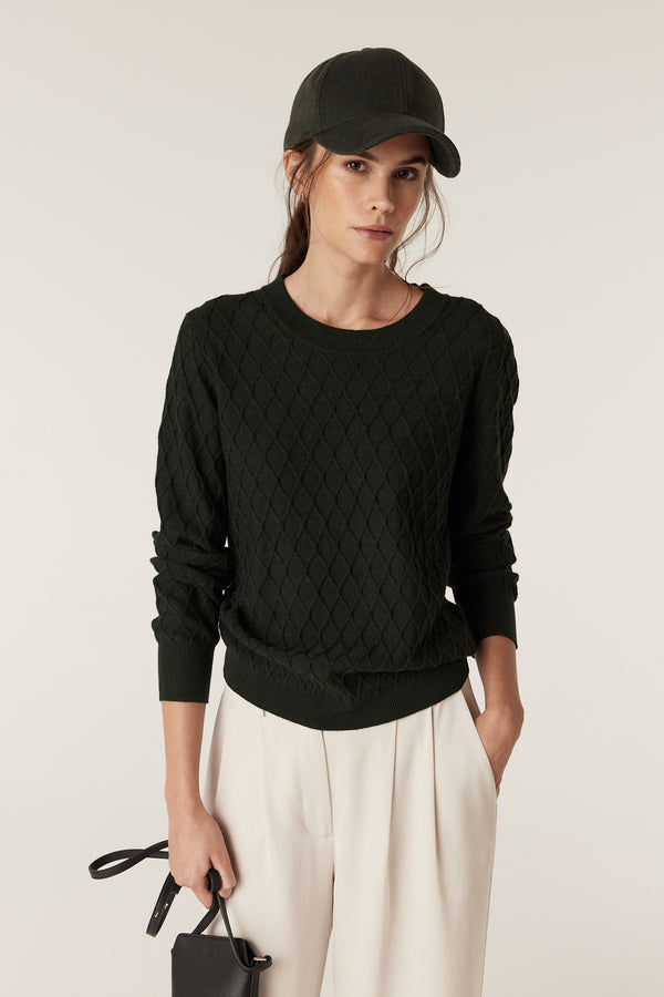 Woman in a moss green Merino wool jumper with diamond cable stitching, cream pants, and a matching cap.