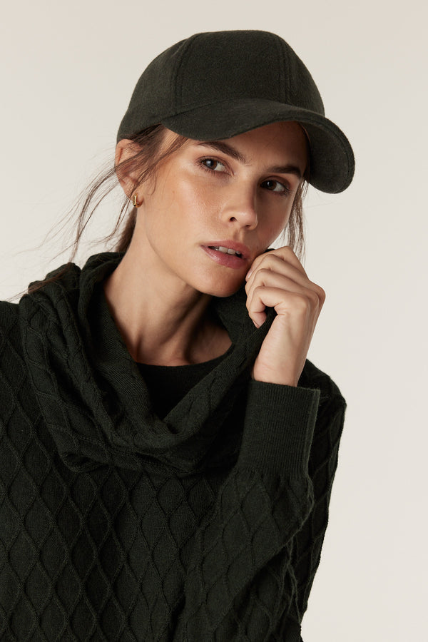 Woman wearing a moss green merino wool snood with diamond cable stitching and a matching baseball cap.