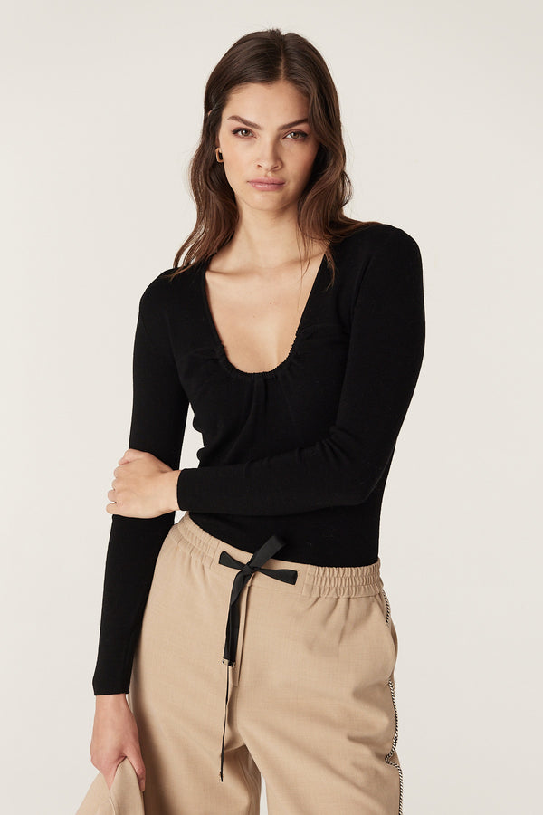 Woman in a black superfine merino gather top with a U-neckline and subtle bust gathers, paired with beige pants.