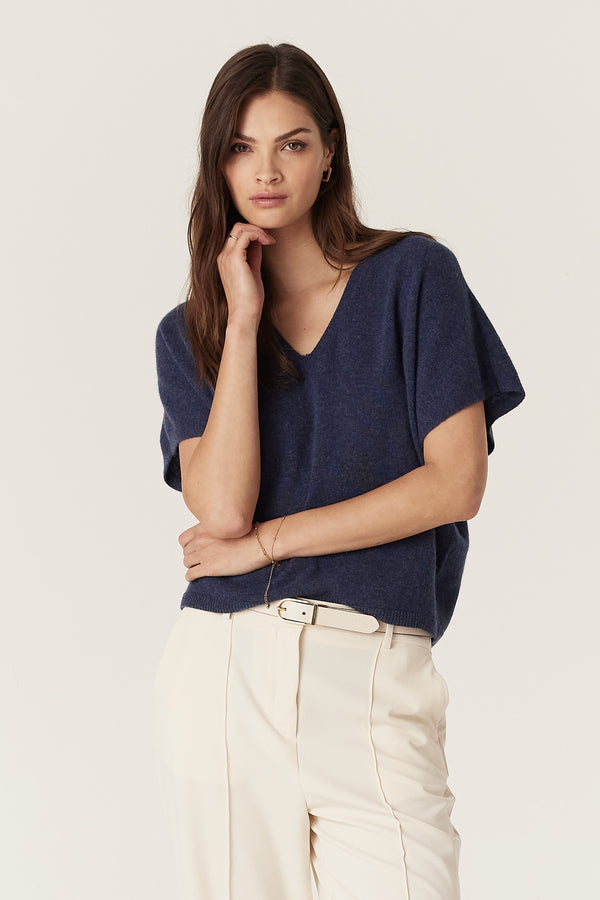 Woman wearing a denim blue cashmere relaxed fit V-neck top with cream pants and belt.