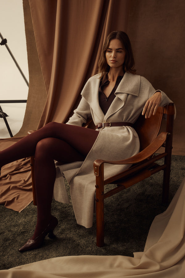 Woman in a mushroom Parsons Merino longline cardigan with a belt, sitting in a leather chair.
