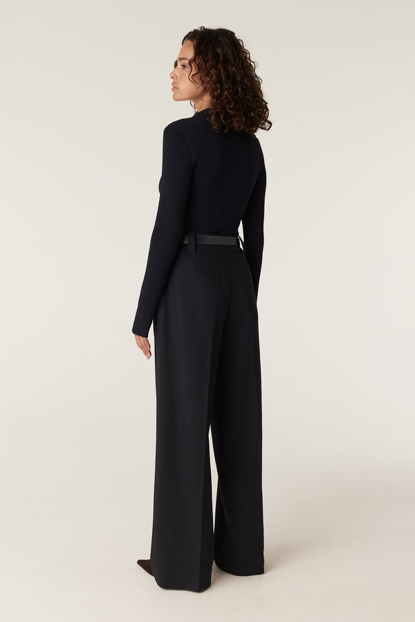 Woman in a navy rib knit polo zip top and wide-leg trousers, viewed from the back.
