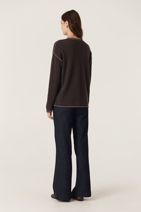 Woman wearing a chocolate pure cashmere V-neck jumper with exposed seams and contrast stitching, paired with dark jeans.