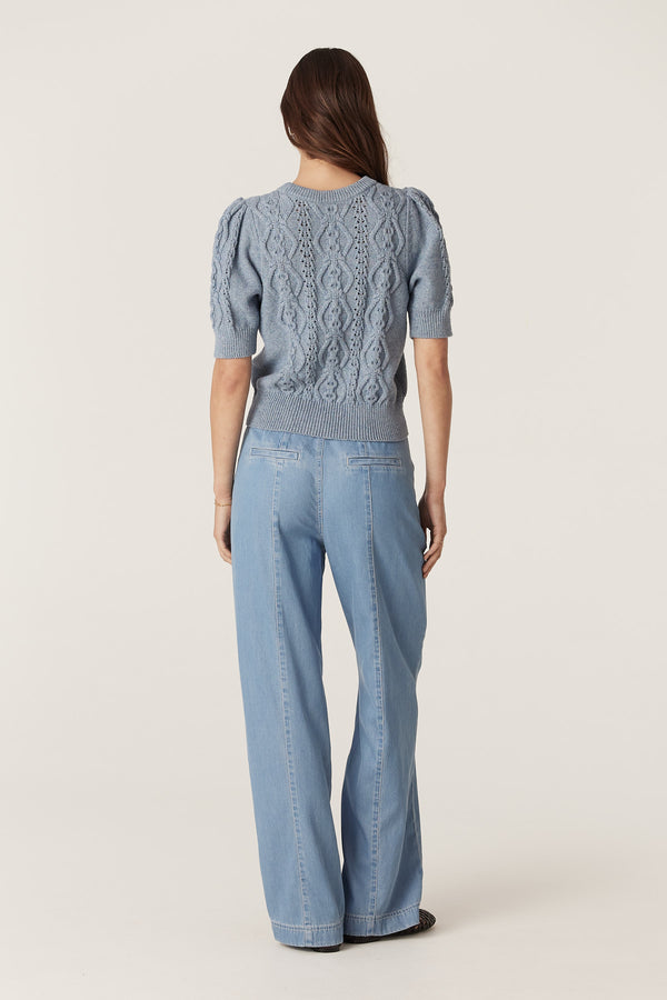 Woman from behind wearing a dusty blue Tavi Knit Top with cable stitch and puffed sleeves, paired with light blue pants.