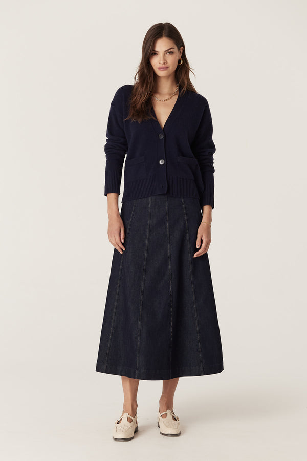 Woman wearing a navy Cashwool V-neck cardigan with front buttons, pockets, and rib trims, paired with a dark denim skirt.