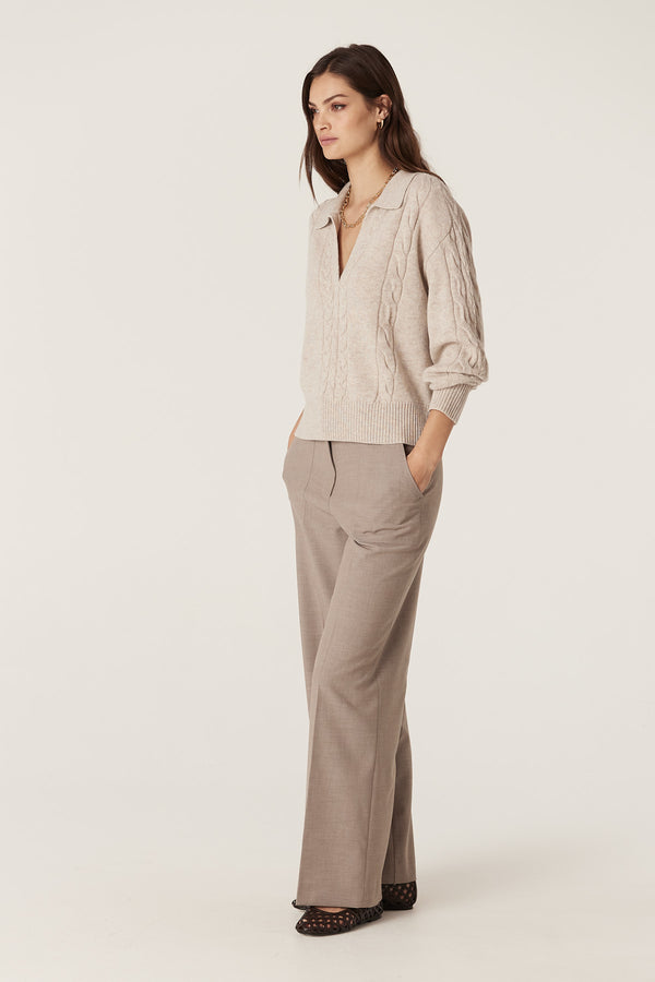 Woman in a sand Cashwool cable knit polo jumper with a V-neck and ribbed trims, paired with brown trousers.