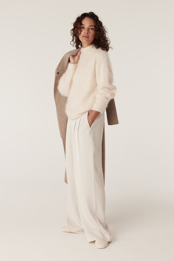 Model in a parchment brushed cashwool turtle jumper, cream wide-leg pants, and a tan coat over her shoulder.