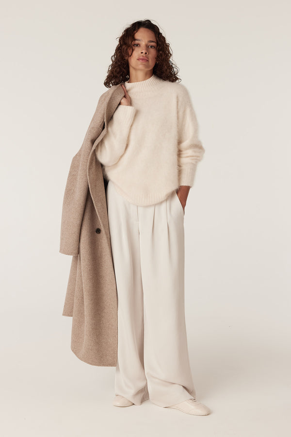 Model in a parchment brushed cashwool turtle neck jumper, cream wide-leg pants, and a tan coat.