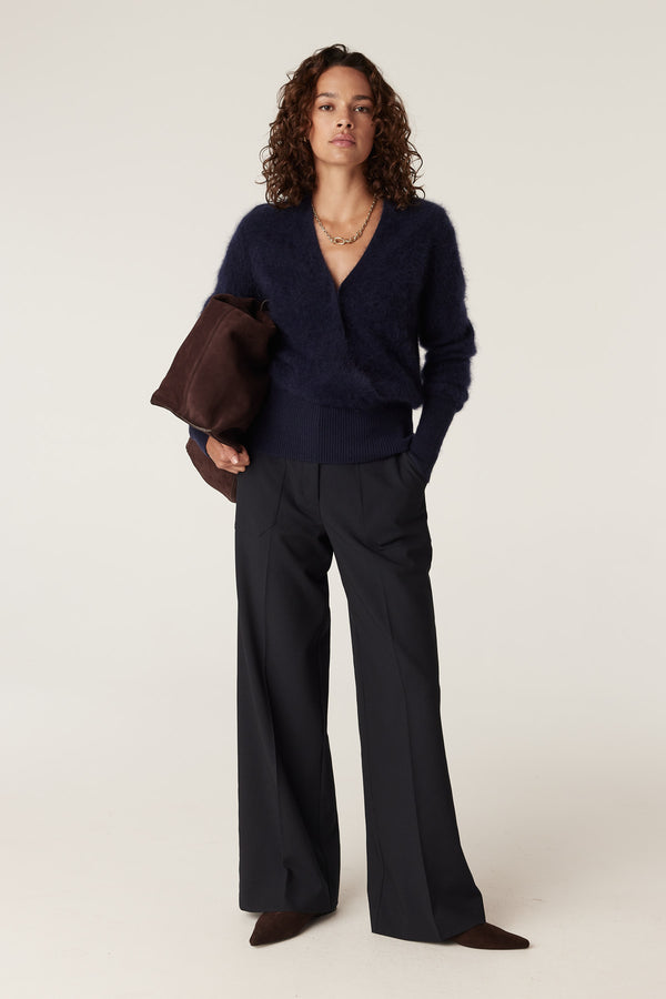 Model wearing a navy brushed cashwool v-neck jumper, dark trousers, and holding a brown suede bag.