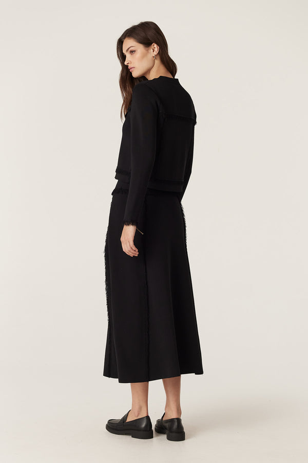 Woman in a black crepe fringe jacket and matching skirt, featuring a crew neck and fringe details.