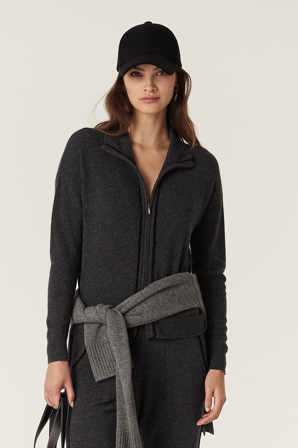 Woman in charcoal Cashwool track zip jumper, tied sweater, and black cap.