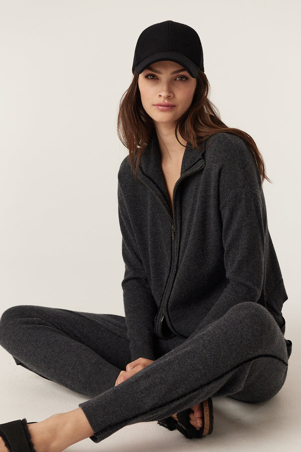Woman in charcoal Cashwool knit track suit, zip-up top, track pants with cord detail, black cap, and sandals.