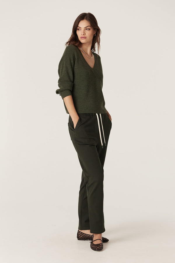 Woman in a forest green Merino wool V-neck faux wrap jumper with statement cuff buttons, dark trousers, and patterned flats.