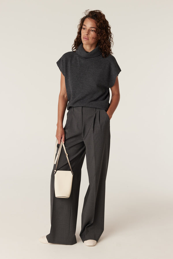 Woman wearing a charcoal merino wool sleeveless turtleneck top with wide-leg trousers and a cream crossbody bag.