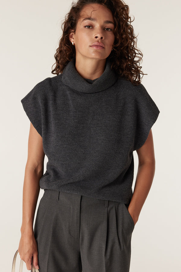 Woman wearing a charcoal merino wool sleeveless top with a cowl neck and dark gray pleated trousers.