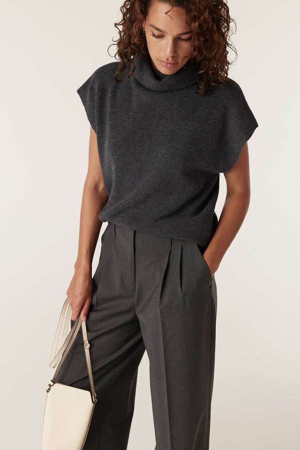 Woman wearing a charcoal merino wool sleeveless turtleneck top and dark grey pleated trousers, holding a cream bag.