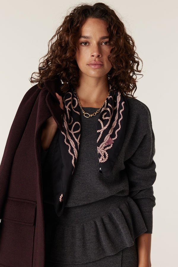 Woman in charcoal merino peplum jumper, holding a burgundy coat, wearing a gold chain and patterned scarf.