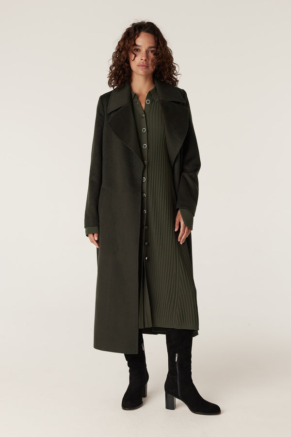 Woman in a long khaki crepe dress with a ribbed skirt, layered under a matching long coat and black boots.