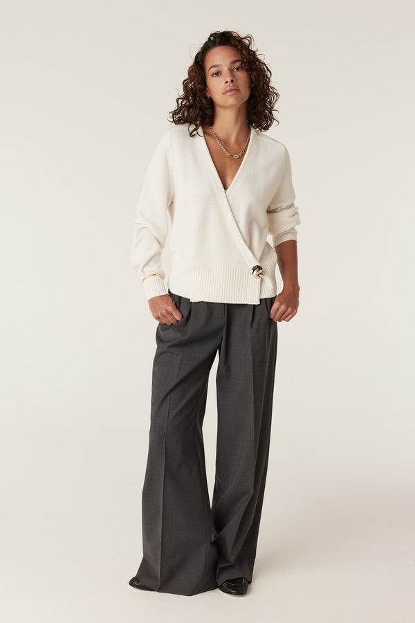 Woman in a milk-colored Cashwool wrap knit jumper with a gold button and dark gray wide-leg trousers.