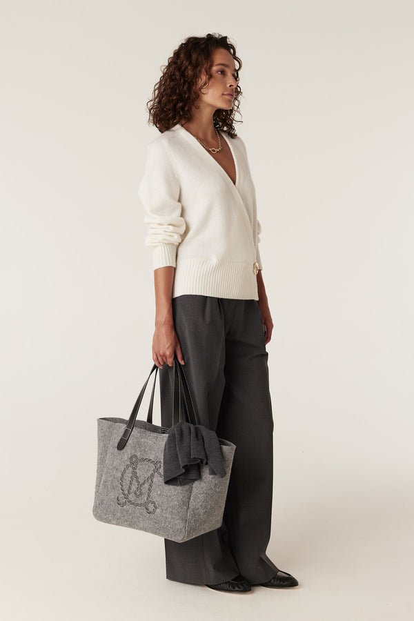 Woman wearing a milk white Cashwool wrap knit jumper, dark grey wide-leg trousers, and holding a grey felt tote bag.