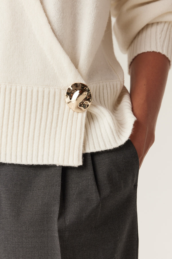 Milk Cashwool wrap knit jumper with a gold button detail, worn over dark gray trousers.