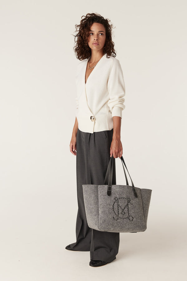 Woman in a milk Cashwool wrap knit jumper with gold button, grey wide-leg pants, and a grey felt tote bag.