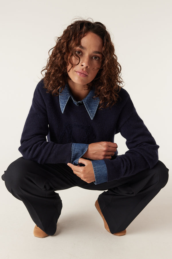 Woman wearing a navy Cashwool logo jumper over a denim shirt, paired with black pants.