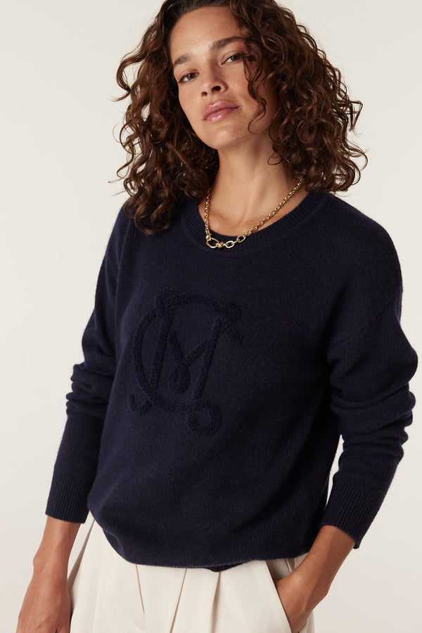 Woman wearing a navy Cashwool logo jumper with an embroidered "M" design, paired with cream pants.