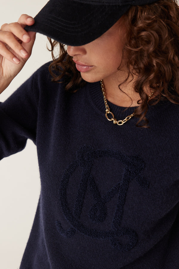 Woman in a navy Cashwool logo jumper with an embroidered "M" and a gold chain necklace.