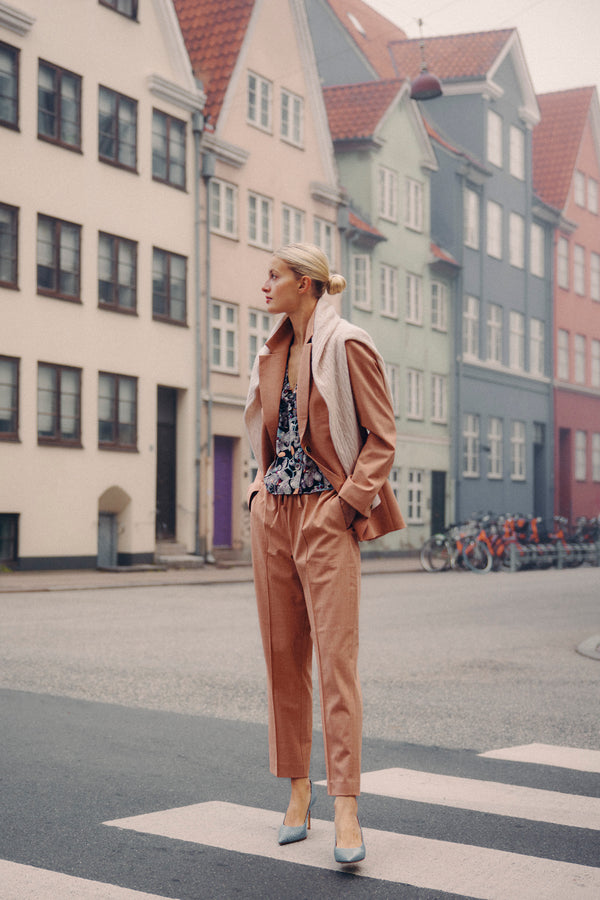 Woman in clay Owen Pant and matching blazer, floral top, and blue heels, standing on a city street.