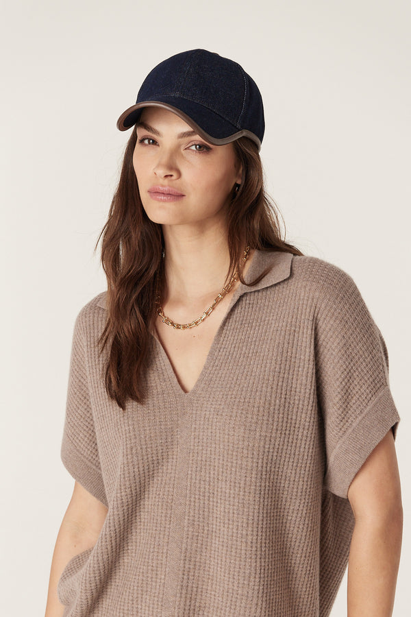 Woman wearing an indigo denim cap with contrast stitching and faux leather trim, paired with a beige knit top.