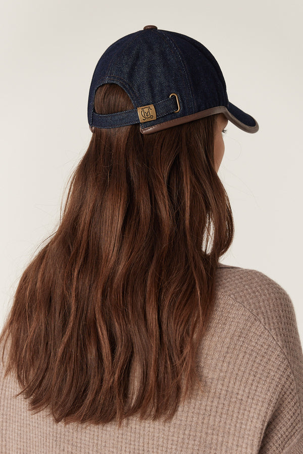 Indigo denim cap with brown faux leather trim, adjustable back strap, and gold slider hardware.
