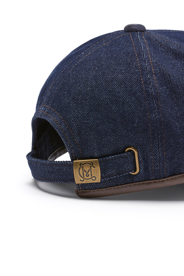 Indigo denim 6-panel cap with contrast stitching, brown faux leather trim, and a brass adjustable slider.
