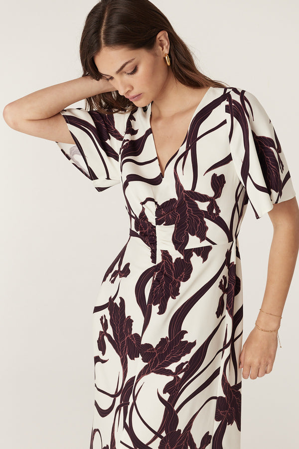 Woman in a white Neve dress with an iris print, V-neck, flared sleeves, and front waist gathers.