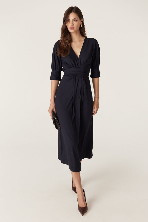 Woman in a navy satin back viscose Holly Dress with V-neck, wrap waist ties, and elbow sleeves.
