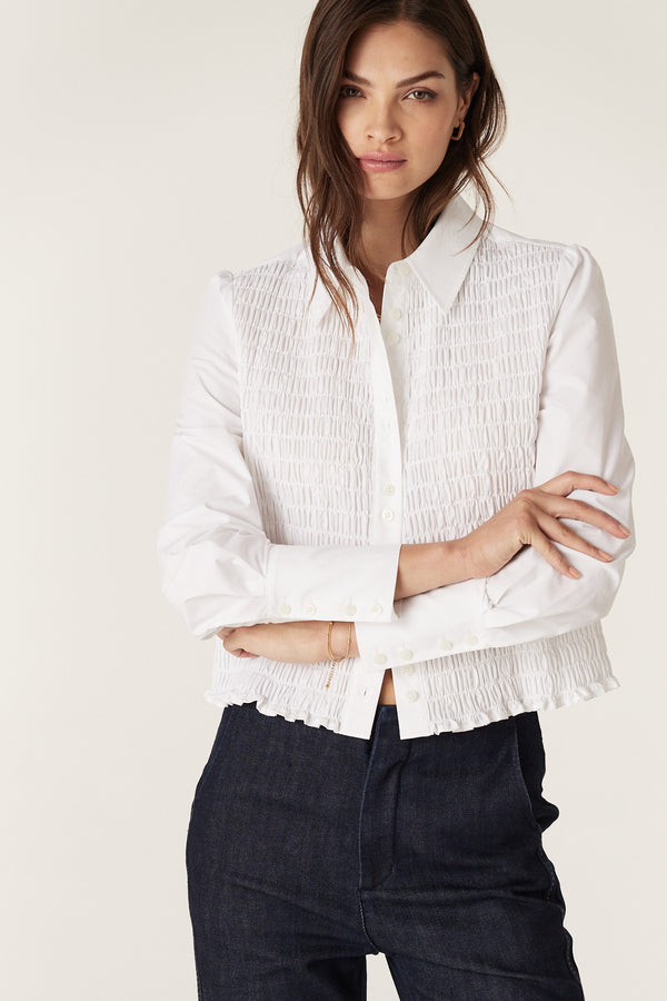 Woman in a white crisp cotton May Shirt with shirred front, long sleeves, and structured cuffs, paired with dark jeans.