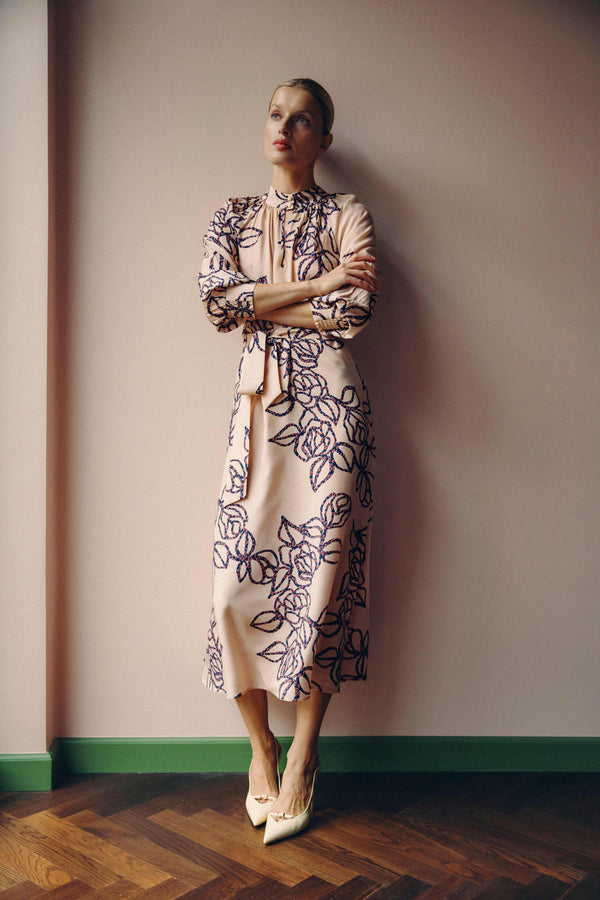 Bloom Dress: Model wears a relaxed fit, pink silk blend dress with blue stencil floral print, stand collar, and waist tie.