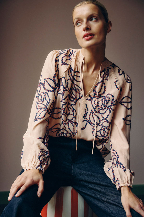 Model wears a Bloom Blouse: a relaxed fit, light pink silk blend blouse with a blue stencil floral print and tie neckline.