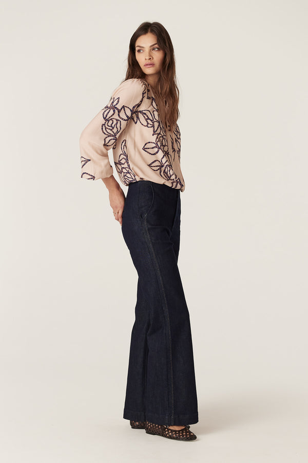 Model wearing a relaxed fit Bloom Blouse in blush silk blend with purple stencil floral print and dark wash jeans.