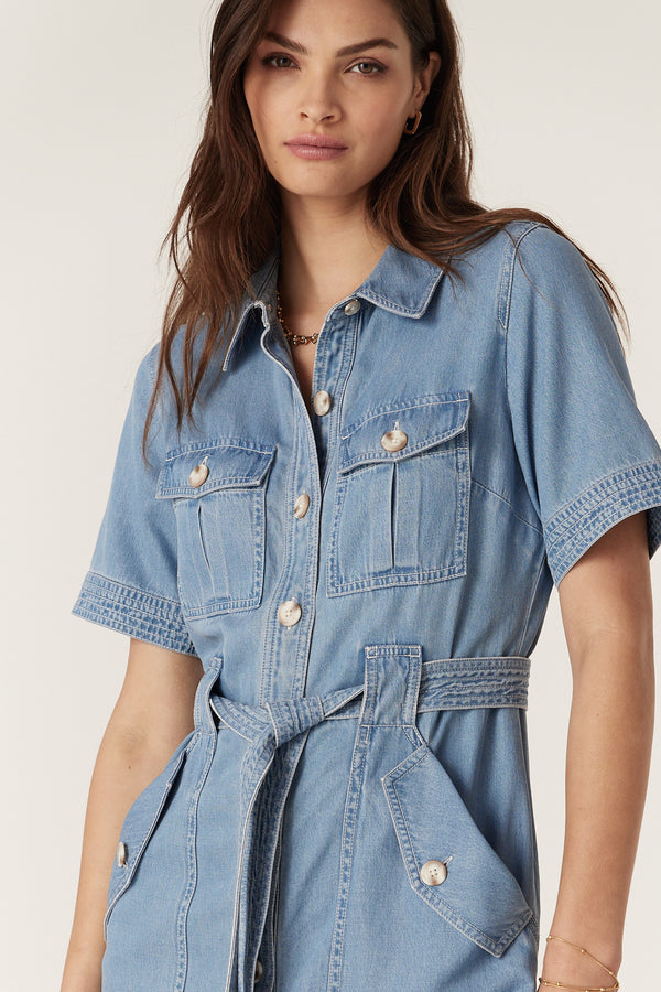 Woman wearing a blue lyocell cotton blend Pearce dress with short sleeves, button front, utility pockets, and waist tie.