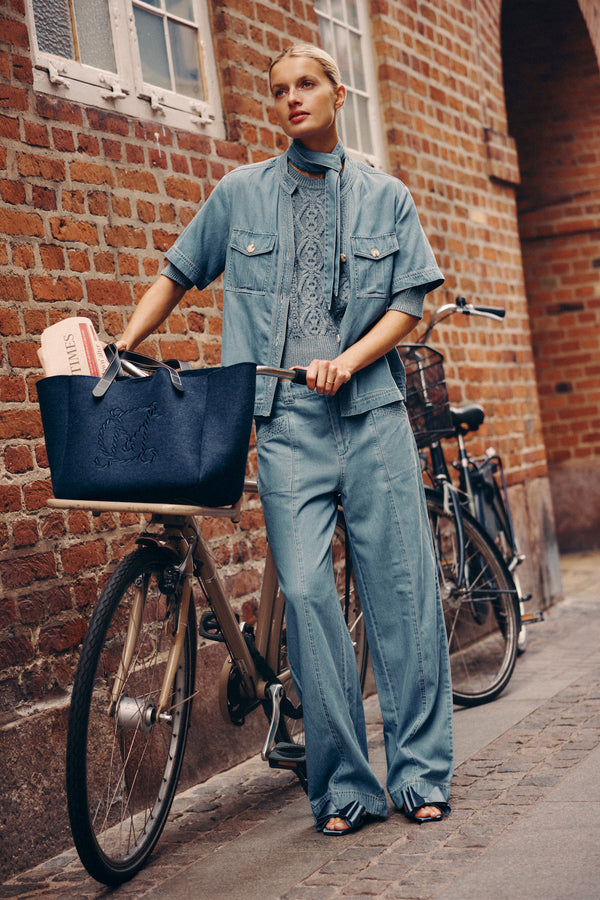 Woman in blue Pearce Pant and matching top, standing with a bike and navy tote bag.