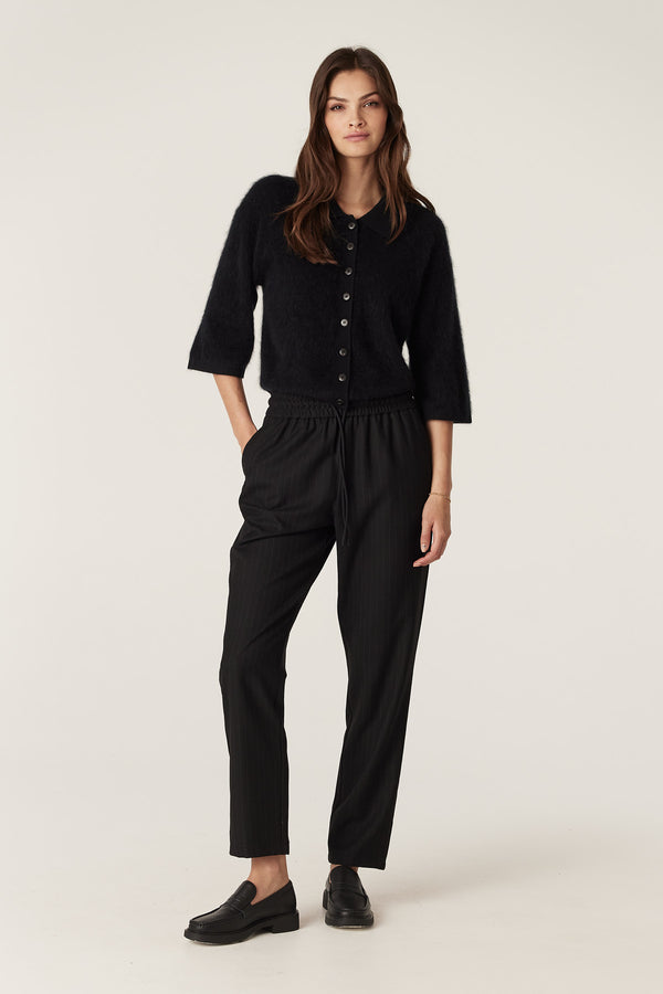 Woman wearing black pinstripe wool blend tapered pants with elastic waist and drawcord, paired with a black cardigan.