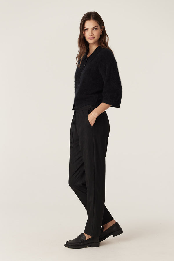 Model wearing black pinstripe Vincent wool blend tapered pants with elasticated waist and front pockets.
