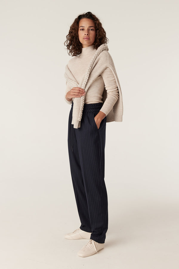 Woman wearing navy pinstripe Vincent wool pants with an elasticated waist and tapered leg, paired with a cream sweater.