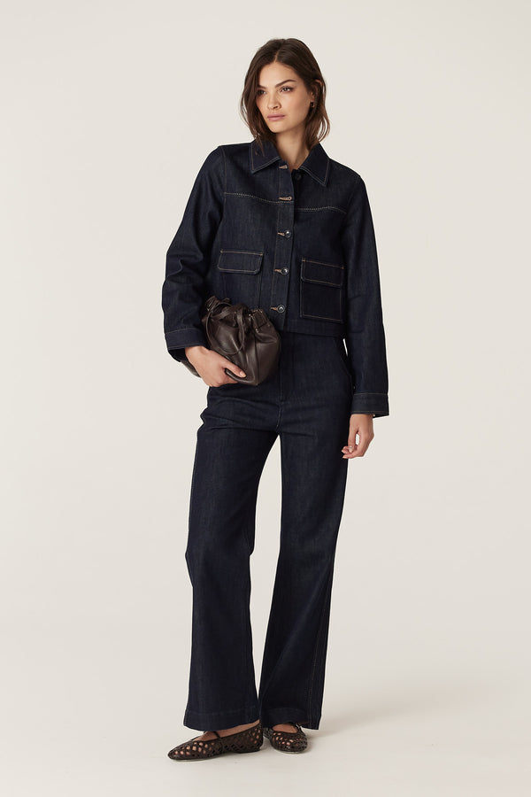 Woman wearing a dark indigo Burke denim jacket and matching jeans, holding a brown leather bag.