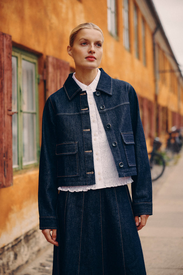 Woman in a dark indigo Burke denim jacket with contrast stitching, white shirt, and matching denim skirt.