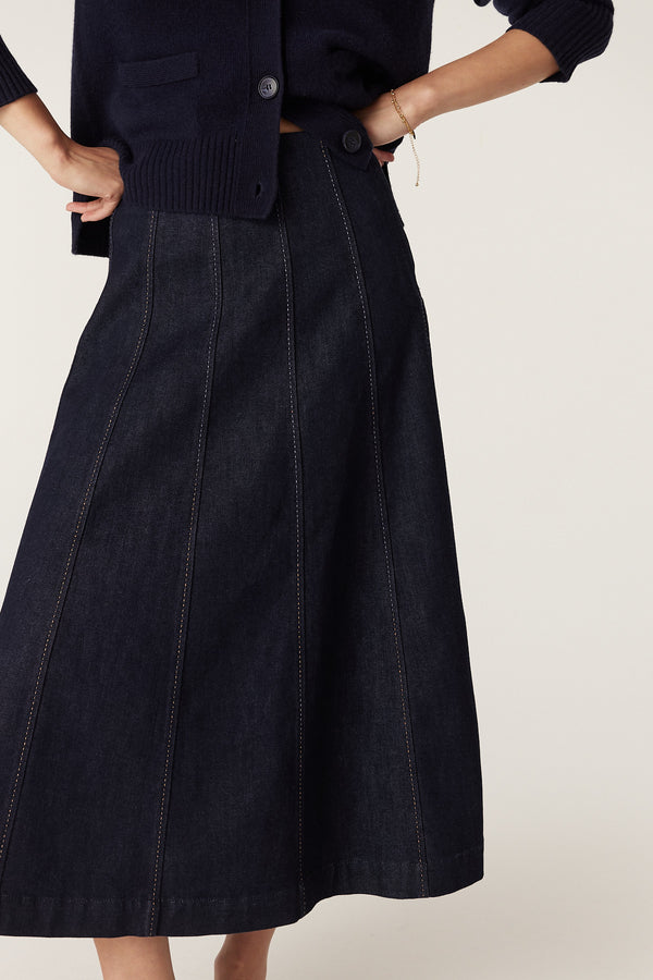 Dark indigo denim midi skirt with gored panels and contrast pick stitching, worn with a navy cardigan.