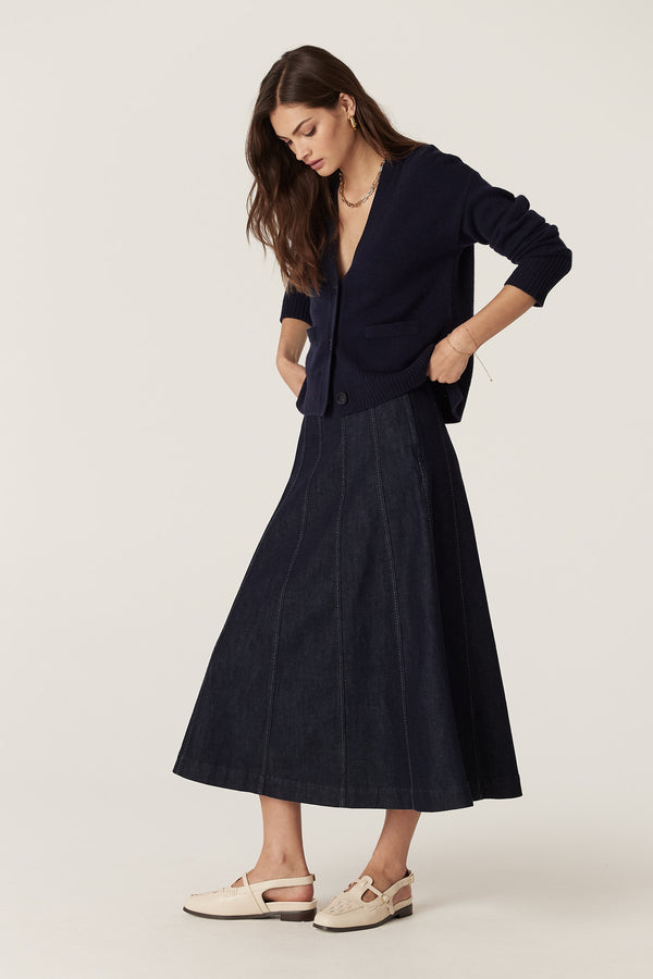 Model wearing a dark indigo Burke denim skirt with gored panels and contrast stitching, paired with a navy cardigan.